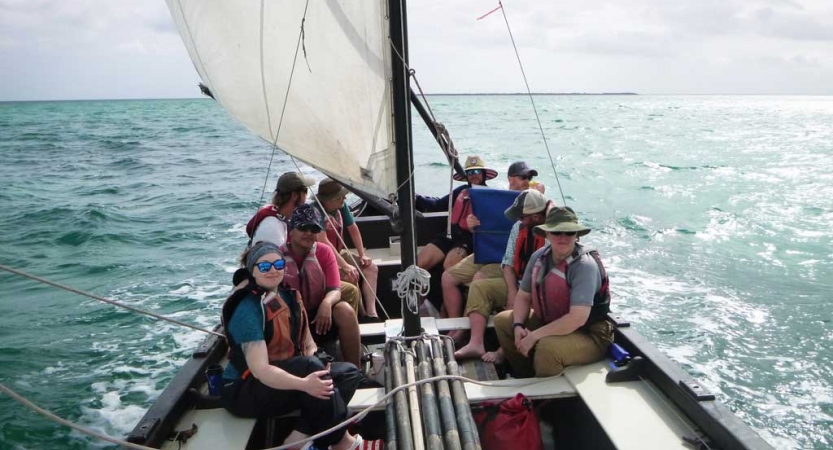 A group of people wearing life jackets sit on a sailboat on an outward bound expedition.